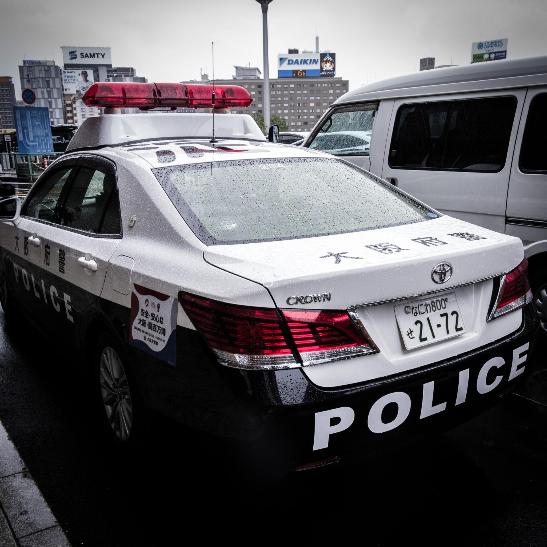 A black and white Japanese police car with a red light bar on top is parked in a city on a rainy day. The word POLICE is written in large letters on the back, and buildings are visible in the background.