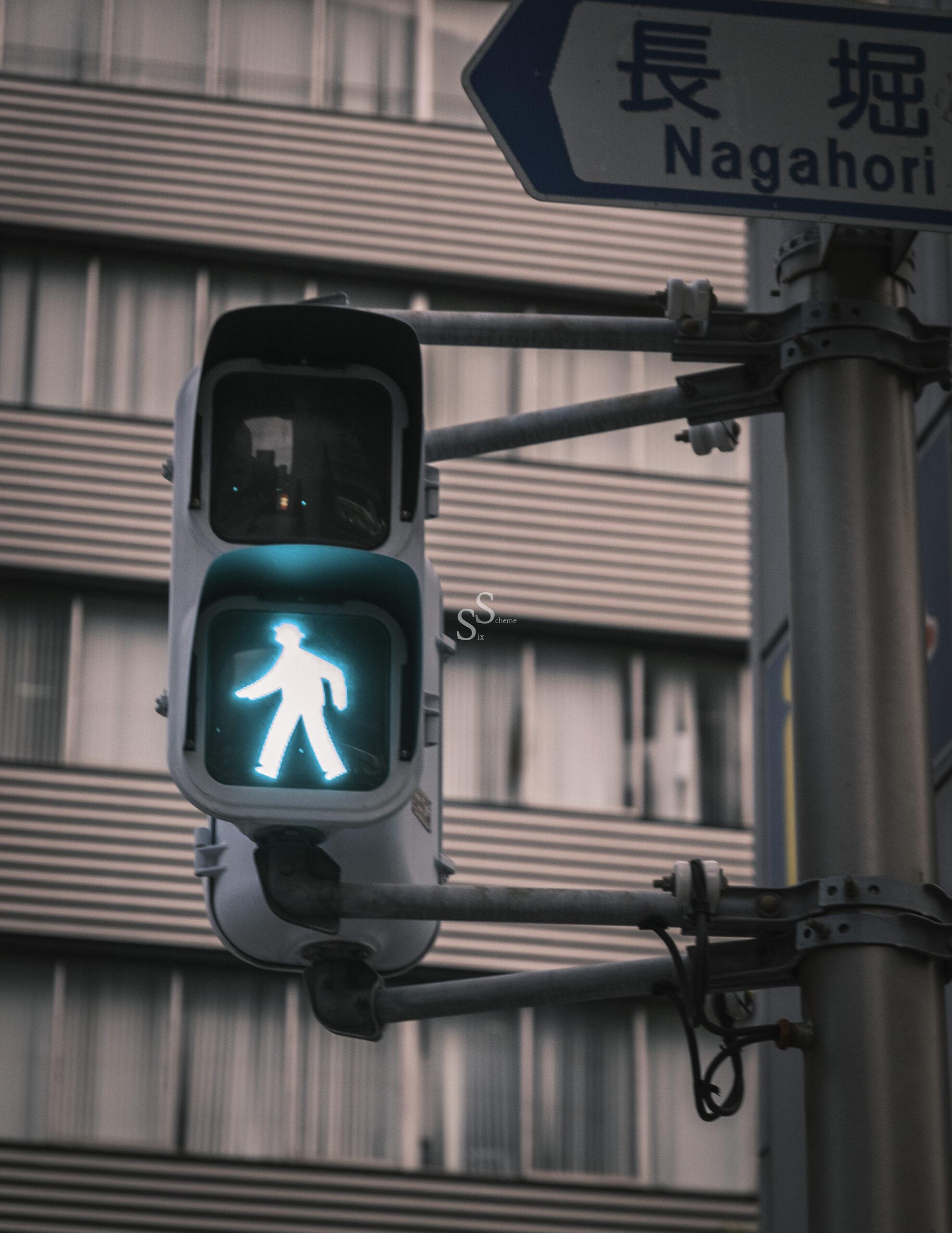 A lit green pedestrian crossing signal in front of a modern building, with a street sign above displaying Japanese characters and the word Nagahori.