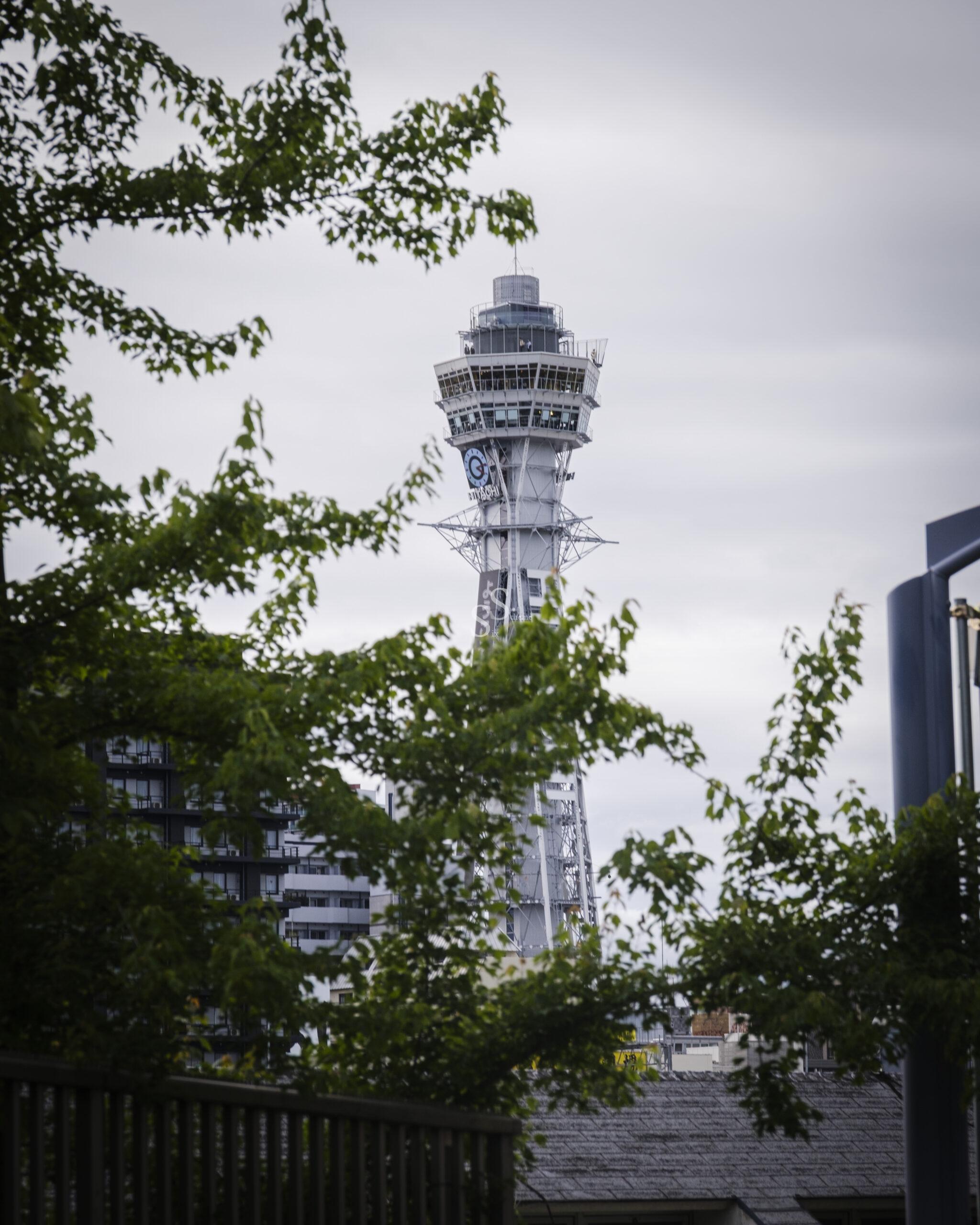 A lofty observation tower, complete with an open-air deck and a sign bearing Japanese writing, stands prominent above rooftops and verdant trees on a cloudy day.