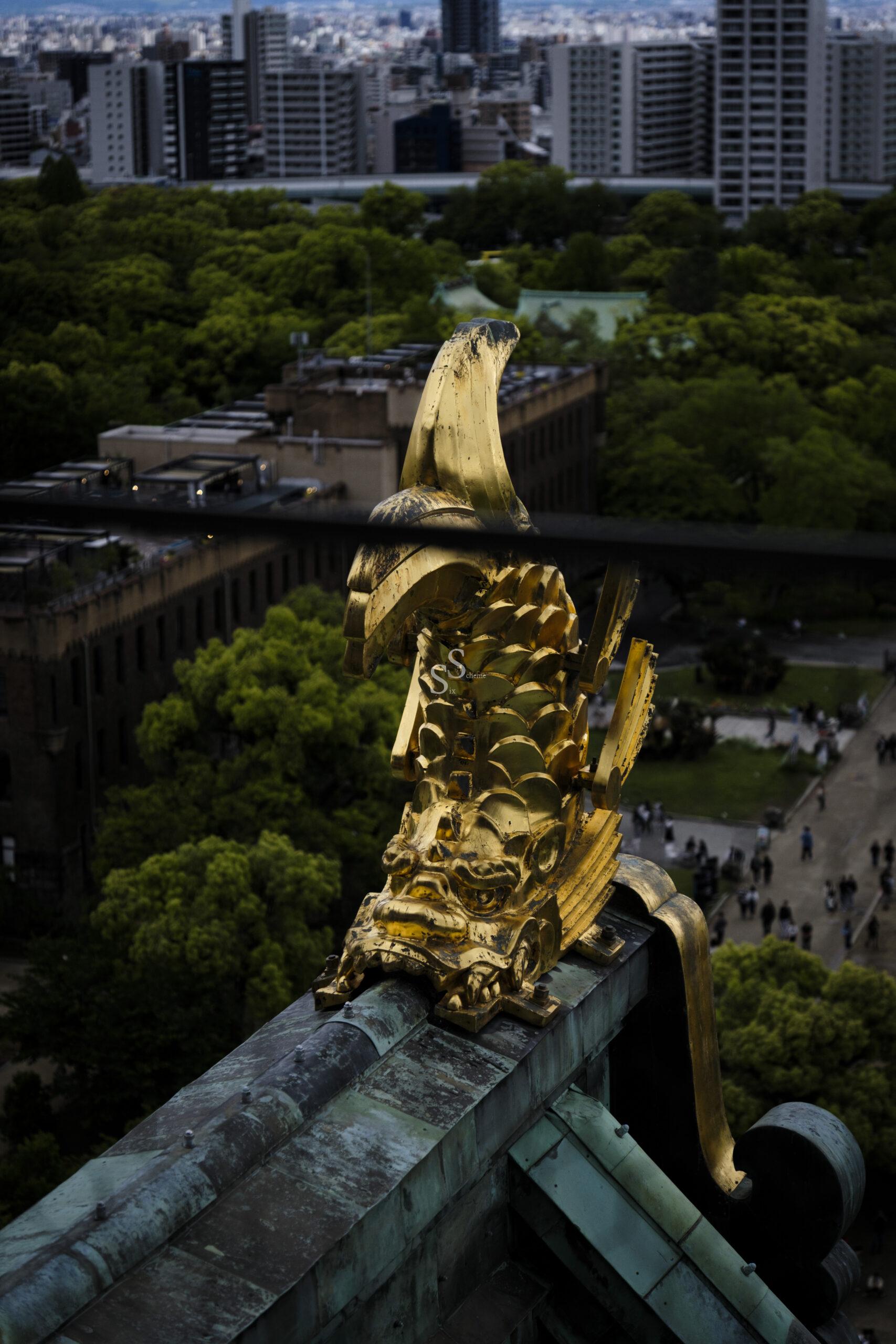 Perched on a rooftop, a golden, fish-shaped ornament gazes out over a city park, its green trees and distant buildings forming the backdrop.