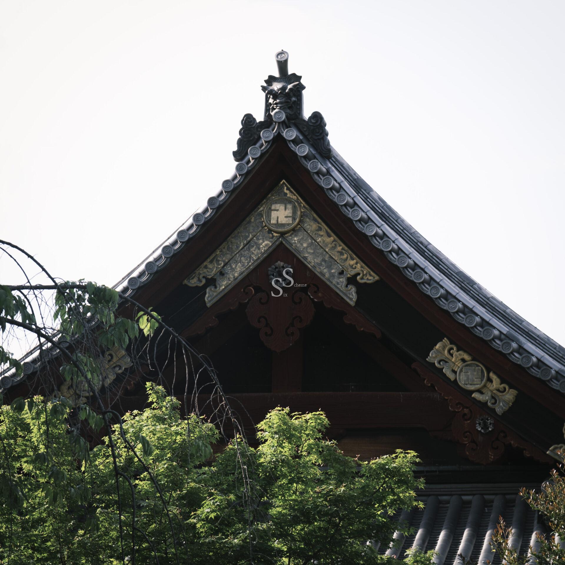 The ornate rooftop of a traditional Japanese building, decorated with intricate carvings and a Manji symbol, rises above green trees against a pale sky.