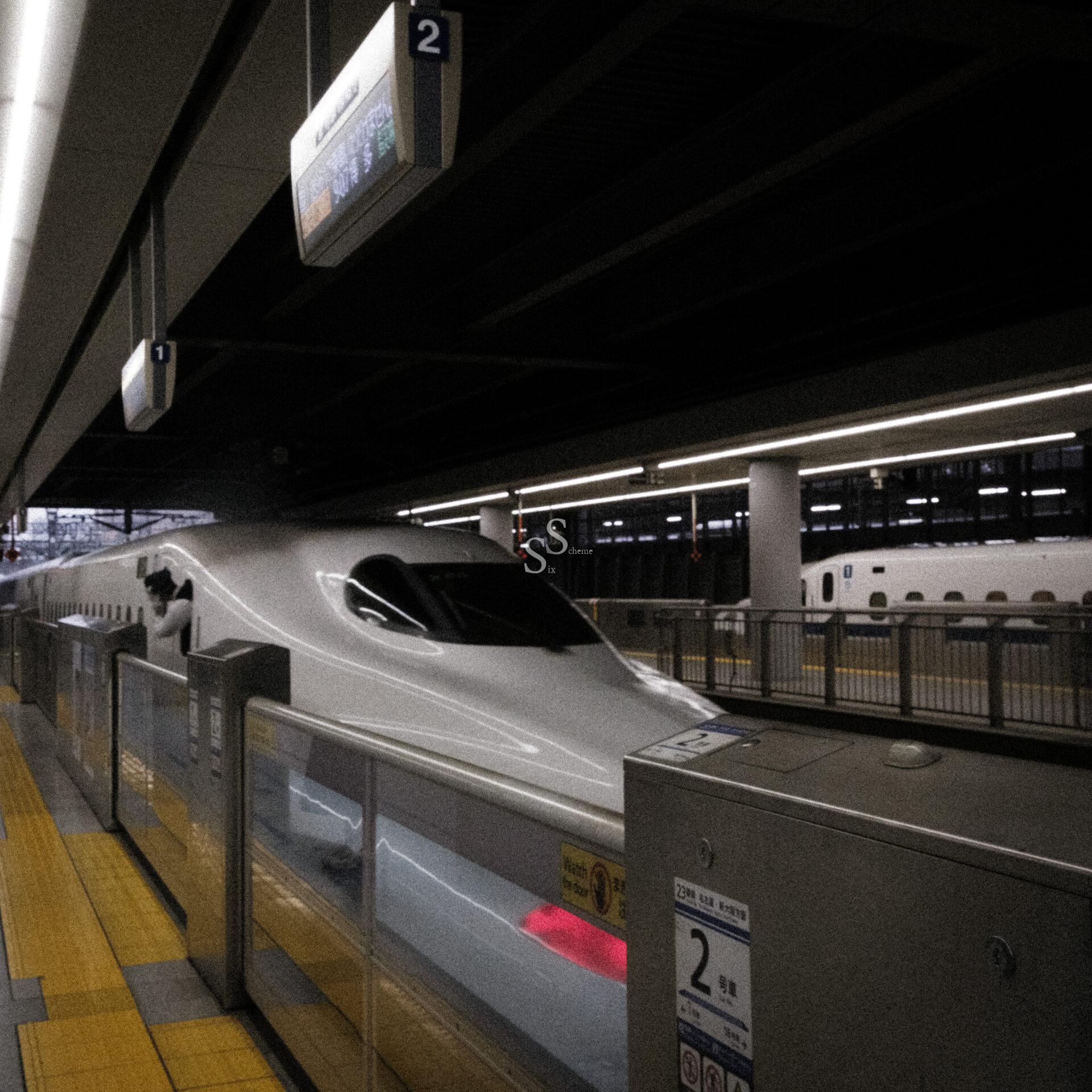 A sleek, white bullet train is stopped at a modern station platform with yellow tactile paving. Overhead signs display the number 2, and another train is visible on a parallel track in the background.