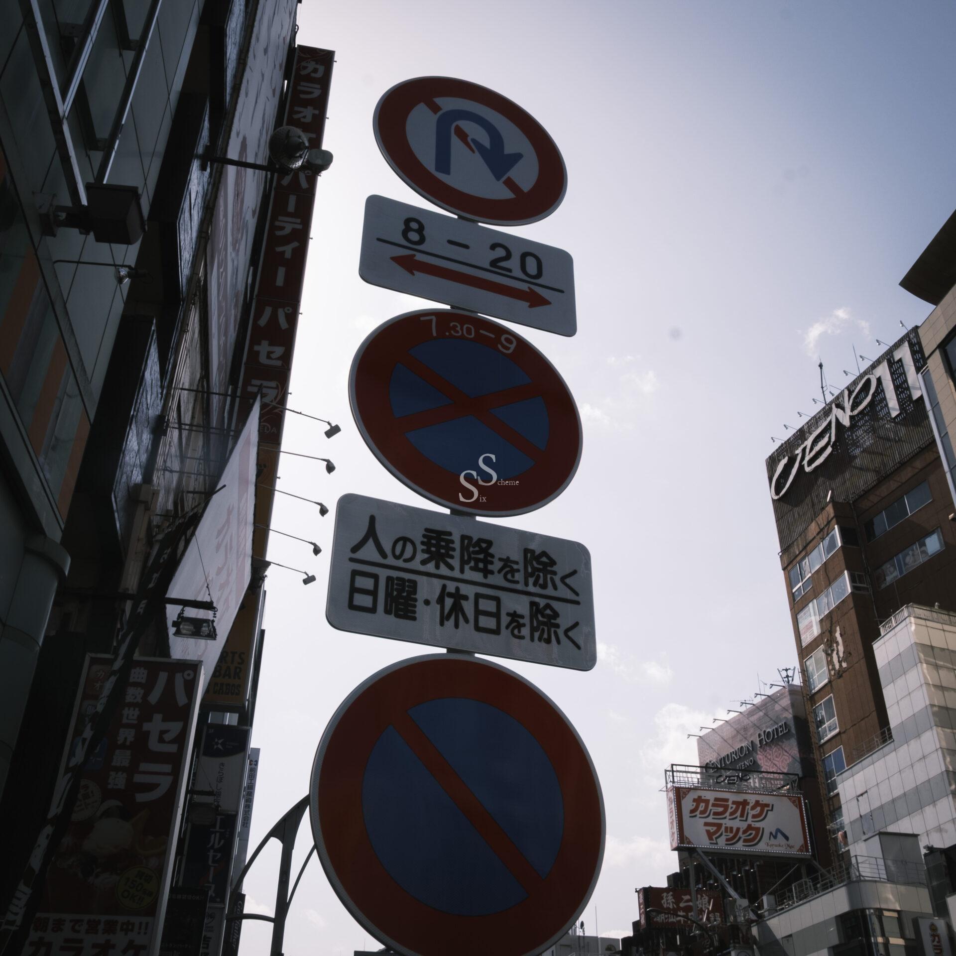 Street signs in Japanese with red and blue symbols, including no parking and no stopping, stand against tall city buildings under a bright sky. Additional signs note exceptions for certain days and drop-offs.