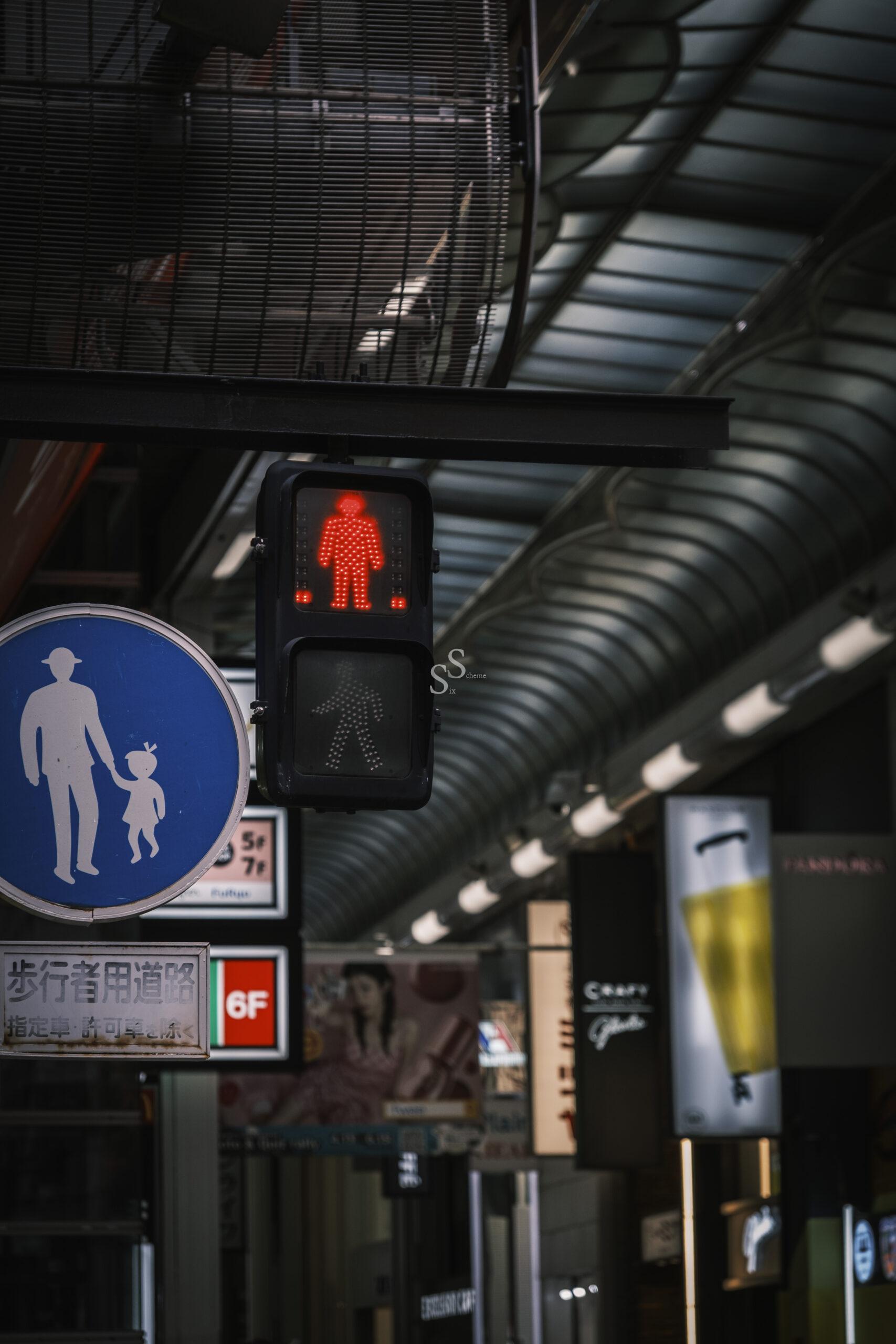 A red pedestrian signal shows a standing figure, indicating not to walk. Below it is a dimmed green signal. Nearby, a blue sign depicts an adult holding a childs hand. The background features illuminated shop signs.