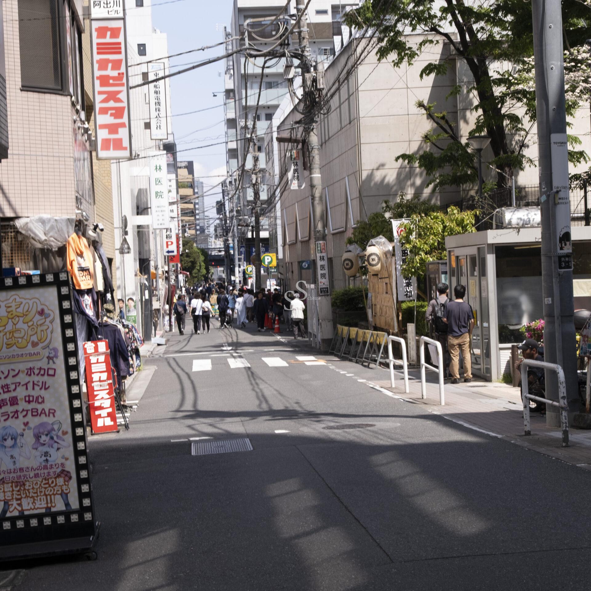 A bustling city street in Japan with people walking on the sidewalk, various signs in Japanese, power lines overhead, and buildings lining both sides of the street.