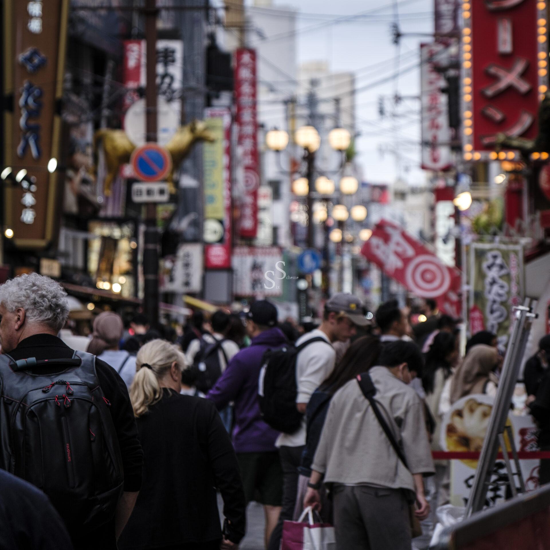 A busy street scene in Japan with many people walking among colorful signs and banners in Japanese, including large red ramen signs. The atmosphere is lively and crowded.