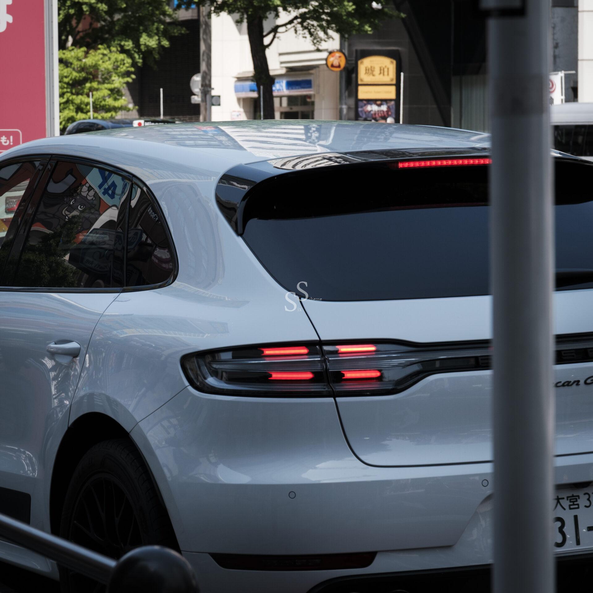 A white SUV with sleek taillights and tinted windows is stopped on a city street, with reflections of buildings and trees visible on the car. A pole partially obscures the view in the foreground.